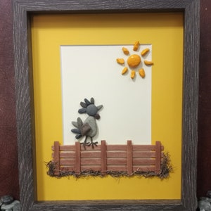May include: A framed artwork featuring a rooster made of stones, standing on a wooden fence with a yellow sun in the background. The artwork is set against a yellow background and framed in a dark brown frame.