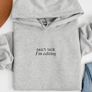 May include: A light gray hoodie with the text "can't talk I'm editing" in black. The hoodie is paired with blue jeans and a brown hat. The sleeves are cuffed to reveal white fabric.