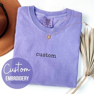 May include: A light purple t-shirt with the word "custom" embroidered on the front. The t-shirt is folded and laid flat on a white surface. The word "Custom Embroidery" is written in a circle on the left side of the image. The word "VIOLET" is written in the bottom left corner of the image.