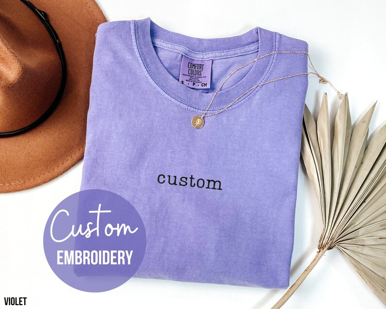 May include: A light purple t-shirt with the word "custom" embroidered on the front. The t-shirt is folded and laid flat on a white surface. The word "Custom Embroidery" is written in a circle on the left side of the image. The word "VIOLET" is written in the bottom left corner of the image.