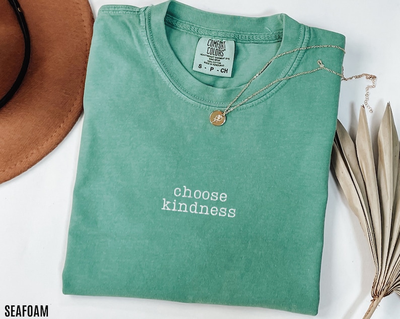 K&ouml;nnte beinhalten: Ein meergr&uuml;nes T-Shirt mit dem Text "choose kindness" in wei&szlig;er Stickerei. Das Shirt ist gefaltet und liegt flach auf einer wei&szlig;en Oberfl&auml;che. Ein brauner Hut und eine goldene Halskette sind im Hintergrund zu sehen.