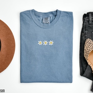 Peut inclure: Un t-shirt bleu jean avec trois marguerites blanches brod&eacute;es. Le t-shirt est pli&eacute; et pos&eacute; &agrave; plat sur une surface blanche.