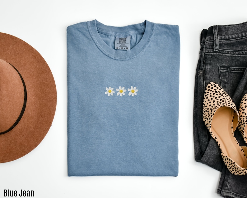 Peut inclure: Un t-shirt bleu jean avec trois marguerites blanches brod&eacute;es. Le t-shirt est pli&eacute; et pos&eacute; &agrave; plat sur une surface blanche.