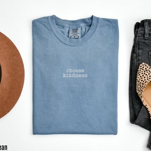K&ouml;nnte beinhalten: Ein blaues Jeans-T-Shirt mit dem Text "choose kindness" in wei&szlig;en Buchstaben. Das Shirt ist gefaltet und liegt flach auf einer wei&szlig;en Oberfl&auml;che. Ein brauner Hut und eine schwarze Jeans mit Leopardenmuster-Flats sind ebenfalls abgebildet.