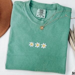 Peut inclure: T-shirt vert d'eau avec un petit motif brod&eacute; de trois marguerites blanches avec des centres jaunes. Le t-shirt a un col rond et est fait d'un tissu doux et confortable. Le nom de la marque "COMFORT COLORS" est visible sur l'&eacute;tiquette.