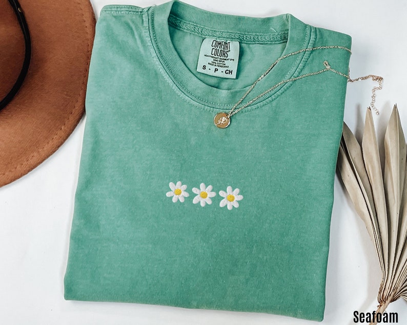 Peut inclure: T-shirt vert d'eau avec un petit motif brod&eacute; de trois marguerites blanches avec des centres jaunes. Le t-shirt a un col rond et est fait d'un tissu doux et confortable. Le nom de la marque "COMFORT COLORS" est visible sur l'&eacute;tiquette.