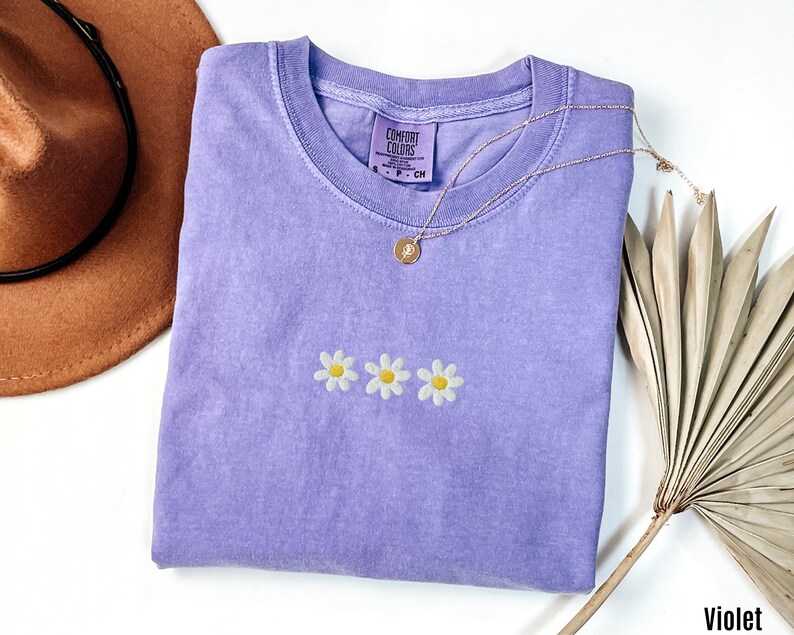Peut inclure: Un t-shirt violet avec un motif de broderie de marguerite. Le t-shirt est pli&eacute; et porte une &eacute;tiquette indiquant "Comfort Colors".
