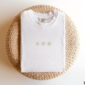 Peut inclure: T-shirt blanc avec trois marguerites brod&eacute;es en jaune et blanc. Le t-shirt est pli&eacute; et pr&eacute;sent&eacute; sur un tapis de paille tress&eacute;.
