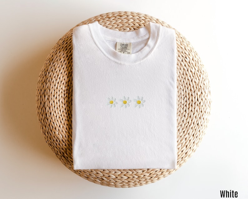 Peut inclure: T-shirt blanc avec trois marguerites brod&eacute;es en jaune et blanc. Le t-shirt est pli&eacute; et pr&eacute;sent&eacute; sur un tapis de paille tress&eacute;.