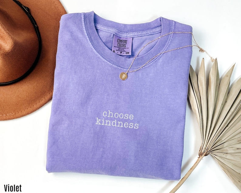 K&ouml;nnte beinhalten: Ein lila T-Shirt mit dem Text "choose kindness" in wei&szlig;en Buchstaben. Das Shirt ist gefaltet und liegt flach auf einer wei&szlig;en Oberfl&auml;che.