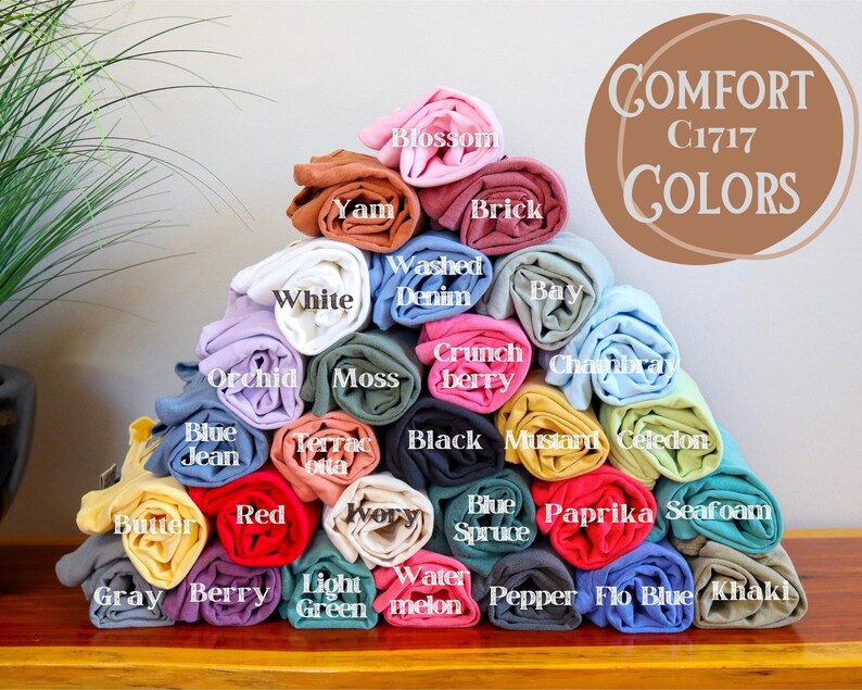 May include: A pyramid of rolled t-shirts in various colors, including red, blue, green, yellow, and pink. The t-shirts are labeled with color names, such as "Berry", "Watermelon", and "Mustard". The text "COMFORT C1717 COLORS" is displayed at the top of the image.