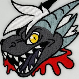 May include: Embroidered patch of a grey and white creature with a large, toothy grin and a red blood splatter. The creature has a yellow eye and a sharp, pointed horn.