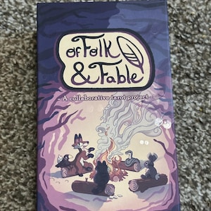 May include: A tarot card box with the title "of Folk & Fable" in a whimsical font. The box features an illustration of woodland creatures gathered around a campfire, with a purple and blue color scheme. The text "A collaborative tarot project" is also visible.