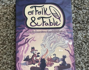 May include: A tarot card box with the title "of Folk & Fable" in a whimsical font. The box features an illustration of woodland creatures gathered around a campfire, with a purple and blue color scheme. The text "A collaborative tarot project" is also visible.