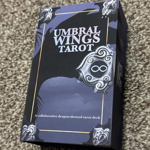 May include: A rectangular tarot card box with the text "UMBRAL WINGS TAROT" in white. The box has a dark blue and black design with gold accents and decorative corner elements. The text "A collaborative dragon-themed tarot deck" is also visible.