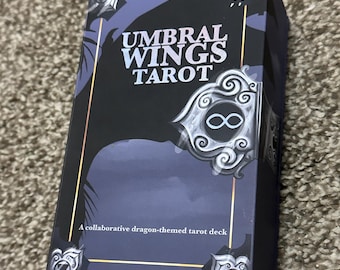 May include: A rectangular tarot card box with the text "UMBRAL WINGS TAROT" in white. The box has a dark blue and black design with gold accents and decorative corner elements. The text "A collaborative dragon-themed tarot deck" is also visible.
