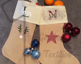 May include: Two beige burlap Christmas stockings with cream-colored cuffs. The left stocking features a green reindeer antler graphic and the name Lars. The right stocking features a red star graphic and the name Nele. Both contain small oranges and ornaments.