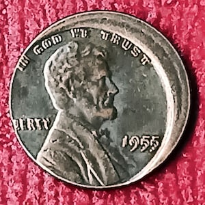 May include: A close-up of a 1955 United States penny. The coin features a profile of Abraham Lincoln, with the inscriptions "IN GOD WE TRUST" and "LIBERTY". The year "1955" is also visible.