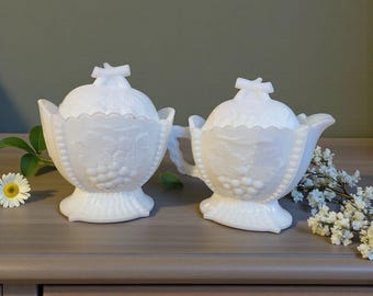 May include: A white milk glass sugar bowl and creamer set. The set has a grape design and beaded trim. The sugar bowl has a lid with a bow-shaped handle. The creamer has a handle and a spout. The set is vintage.