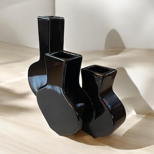 May include: Three black ceramic vases of different sizes, each with a rounded base and a square opening at the top. The vases are arranged in a staggered formation, with the largest vase in the back and the smallest vase in the front.