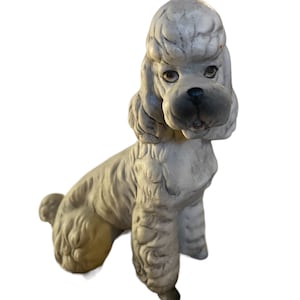 May include: A white ceramic figurine of a poodle dog sitting with its head tilted to the side. The dog has a detailed face with brown eyes and a black nose.