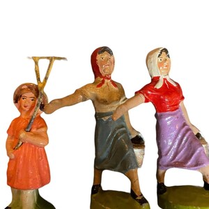 May include: Three hand-painted figurines of women wearing traditional clothing. The woman on the left is holding a rake, the woman in the middle is carrying a bucket, and the woman on the right is holding a bucket. The figurines are standing on green bases.