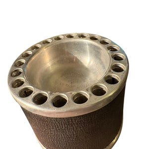 May include: Vintage ashtray with a silver-toned metal top and a dark brown textured base. The top features numerous circular openings around the rim. The ashtray has a cylindrical shape and a classic design.