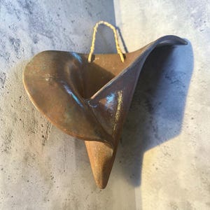 May include: A handcrafted, bronze-colored ceramic wall pocket with a calla lily shape. The vessel has a curved, open design with a textured surface and a woven cord for hanging. The wall pocket is mounted on a gray concrete wall.