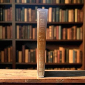 Puede incluir: Un libro de tapa dura titulado "The Hidden Years" con una cubierta de cuero marrón se encuentra sobre una superficie de madera. El libro tiene letras doradas y cuadrados decorativos en relieve. El fondo presenta una estantería borrosa llena de libros.