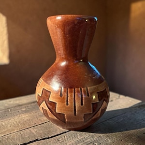 May include: A small, bulbous ceramic vase with a glossy, dark brown finish. The lower section features a lighter tan, geometric pattern carved into the surface, consisting of repeating stepped shapes and vertical lines encircling the base.
