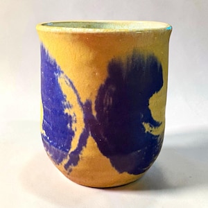 May include: A cylindrical ceramic cup with a matte yellow base and bold, abstract blue brushstroke patterns wrapping around the exterior. The rim is smooth and slightly rounded, featuring a light-colored interior glaze finish.