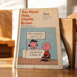 Puede incluir: Una portada de libro vintage de Peanuts con Charlie Brown y Lucy. La portada muestra a Lucy de pie detrás de una mesa con un cartel que dice "Psychiatric Help 5¢". Charlie Brown está sentado en un taburete frente a la mesa, con aspecto triste. El título del libro es "You Need Help, Charlie Brown".