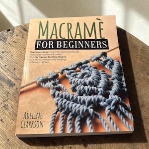 May include: A softcover book titled Macrame For Beginners by Adeline Clarkson. The cover features a light tan background with dark green and white text, and an illustration of blue macrame cord knotted onto a wooden dowel rod.