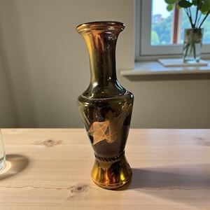 Puede incluir: Un jarrón decorativo de vidrio con un acabado degradado dorado y marrón oscuro. El jarrón presenta una parte superior acampanada y una base bulbosa con un detalle similar a una cuerda. Un diseño geométrico dorado es visible en la parte delantera del jarrón.