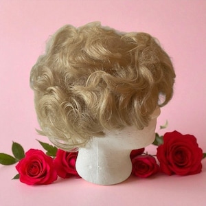 Peut inclure: Une perruque blonde aux boucles douces est présentée sur une tête en polystyrène blanche. La perruque est posée sur un fond rose et entourée de roses rouges vibrantes, créant une esthétique classique et élégante.