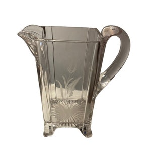 Könnte beinhalten: Klarer Glaskrug mit quadratischem Körper, gebogenem Griff und Ausguss. Der Krug hat geätzte botanische Designs auf der Vorderseite und wird von drei kleinen Füßen getragen. Ein klassisches Stück zum Servieren von Getränken.