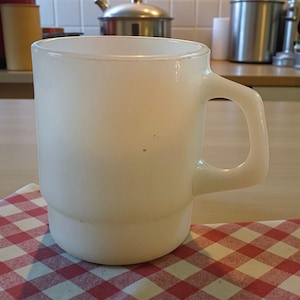 Puede incluir: Una taza vintage de color blanco opaco con un asa redondeada. La taza está colocada sobre un paño a cuadros rojos y blancos. La taza tiene forma cilíndrica con una base ligeramente ensanchada. El fondo incluye elementos de cocina.