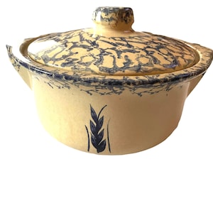 May include: A round, cream-colored ceramic casserole dish with a matching lid. The exterior features a dark blue sponge-painted pattern along the rim and lid, with a single dark blue painted wheat stalk motif centered on the front of the vessel.