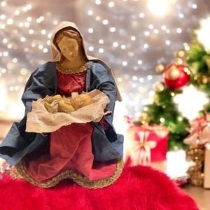 Könnte beinhalten: Eine blaue und rote Figur von Maria, die ein Jesuskind hält. Die Figur sitzt auf einer roten, flauschigen Oberfläche.