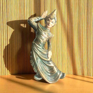 May include: A ceramic figurine of a dancer wearing a pointed headpiece and a long, flowing gown. The figure is finished in a metallic teal and gold glaze, with one arm raised near the head and the other arm positioned at the side of the waist.