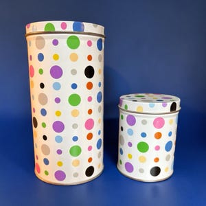 May include: Two cylindrical metal canisters with a white base featuring a colorful pattern of polka dots in various sizes and shades, including green, purple, blue, orange, yellow, pink, grey, and black, with matching lids on each container.