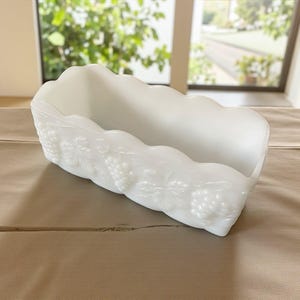May include: A white, rectangular milk glass dish with a scalloped edge. The dish has a raised grape and leaf design on the sides. The dish is empty and rests on a neutral-coloured surface.