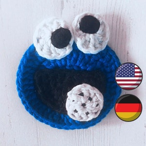 May include: Blue crocheted cookie monster character with white and black eyes and a white crocheted mouth. The character is on a white wooden background. The image also includes the American and German flags.