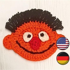 May include: Orange crocheted character with black hair, brown eyes, and a red nose. The character is smiling. The image also includes the American and German flags.