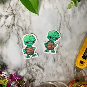 May include: Two green cartoon turtle stickers with brown shells. The turtles have big eyes and are standing on their hind legs.