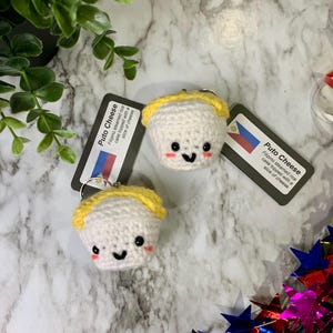 May include: Two crocheted Puto Cheese keychains with yellow and white yarn. The keychains are shaped like a small cup with a smiling face. The text on the card reads "Puto Cheese Filipino steamed rice cake topped with a slice of cheese."