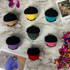 May include: Seven handmade crochet acorn keychains in various colors, including tan, yellow, teal, gray, purple, pink, and red, each with a black cap. The keychains are arranged on a marble surface, with two photos and colorful decorations.