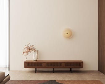 May include: A light wood media console with a slatted design and black metal legs. A white vase with dried branches sits on top of the console. A round, white light fixture is mounted on the wall behind the console.