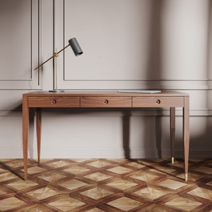 May include: A wooden desk with three drawers and gold-colored legs. The desk is in front of a white wall with a gold-colored lamp on the left side.