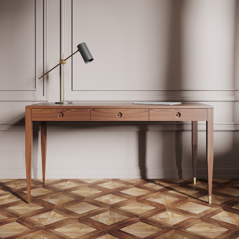 May include: A wooden desk with three drawers and gold-colored legs. The desk is in front of a white wall with a gold-colored lamp on the left side.